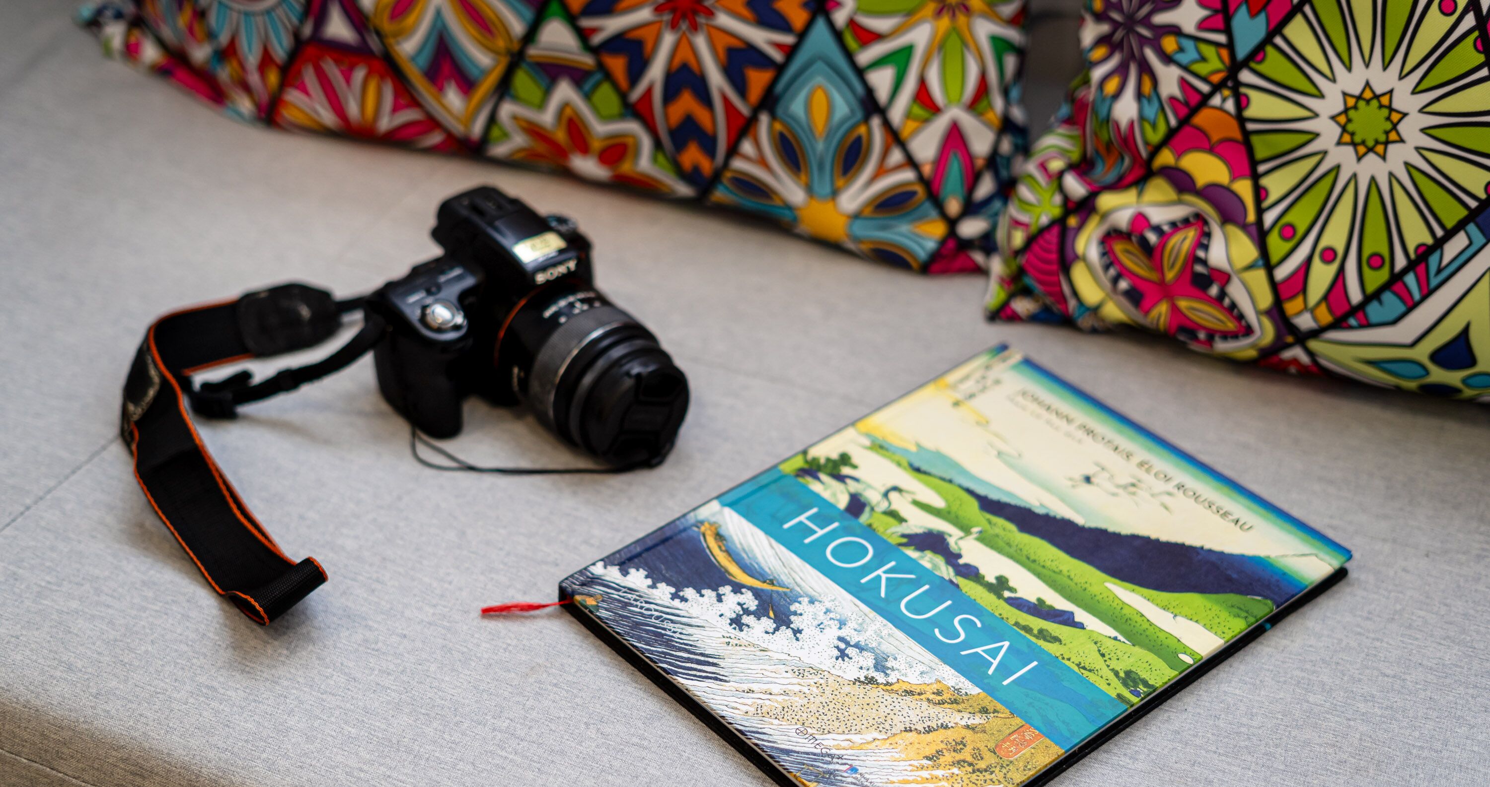 Et kamera og en bog ligger på en overflade med farverige puder med mønster i baggrunden.