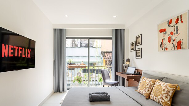 Een moderne hotelkamer met een groot bed, een bureau met een laptop, een televisie waarop 'NETFLIX' wordt weergegeven en een balkon met uitzicht op de stad.