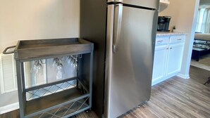 Un refrigerador de acero inoxidable en una cocina con piso de madera, gabinetes blancos y una pequeña repisa con cristalería.