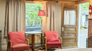 Una habitación con piso de madera, dos sillas tapizadas en rojo, una cama con cubrecama acolchado, una mesa con lámpara y una puerta que da al exterior.