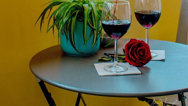 Une table sur laquelle se trouvent un pot de fleurs, une rose rouge et deux verres à vin remplis de vin rouge, et une bouteille dont on verse le contenu dans l’un des verres.