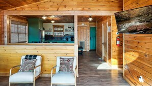 L’intérieur d’un chalet en bois comprend un coin cuisine, un espace détente avec des coussins à motifs et un téléviseur fixé au mur qui diffuse des images de paysages naturels.