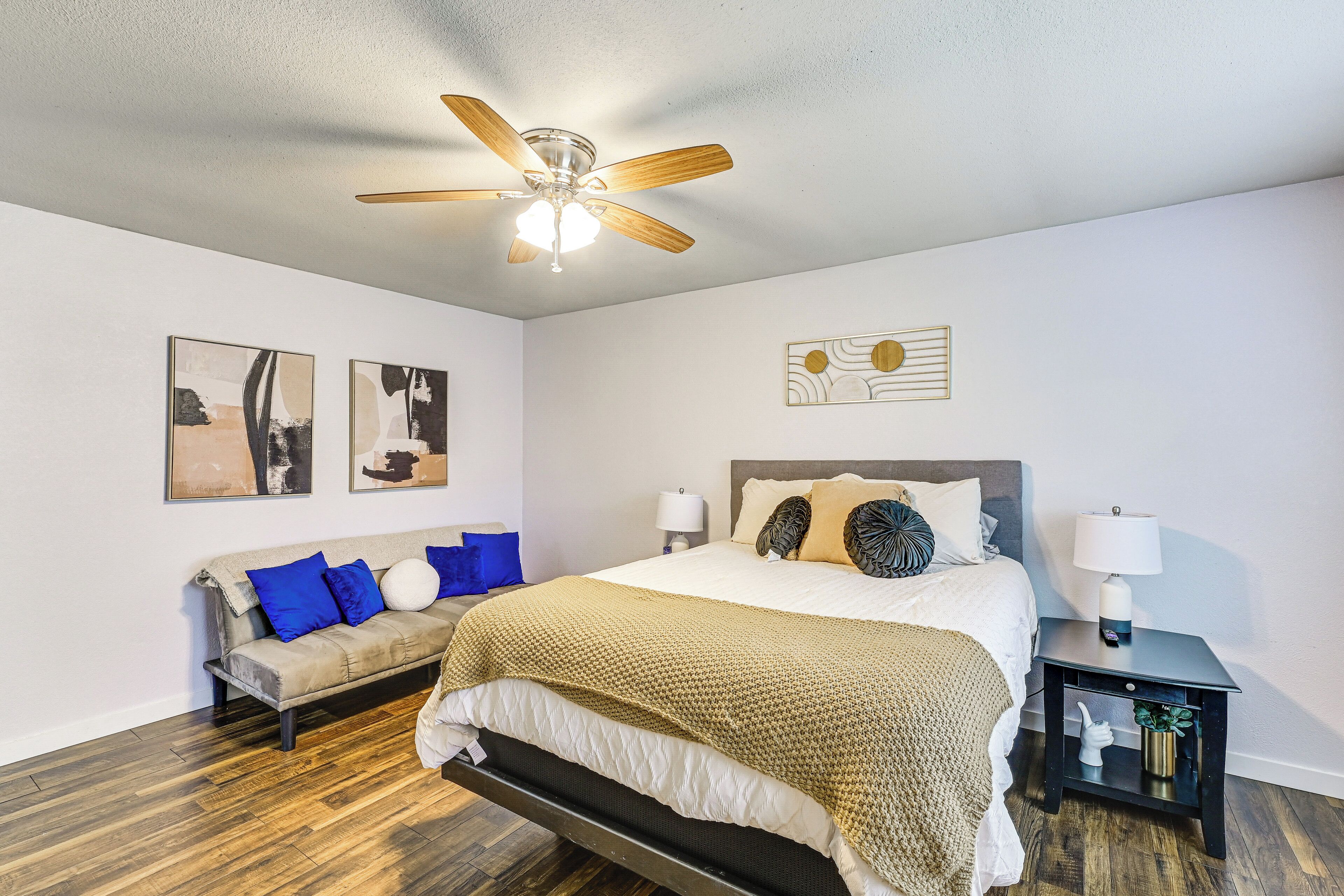 A bedroom with a bed, a nightstand, a lamp, a ceiling fan, and artwork on the walls.