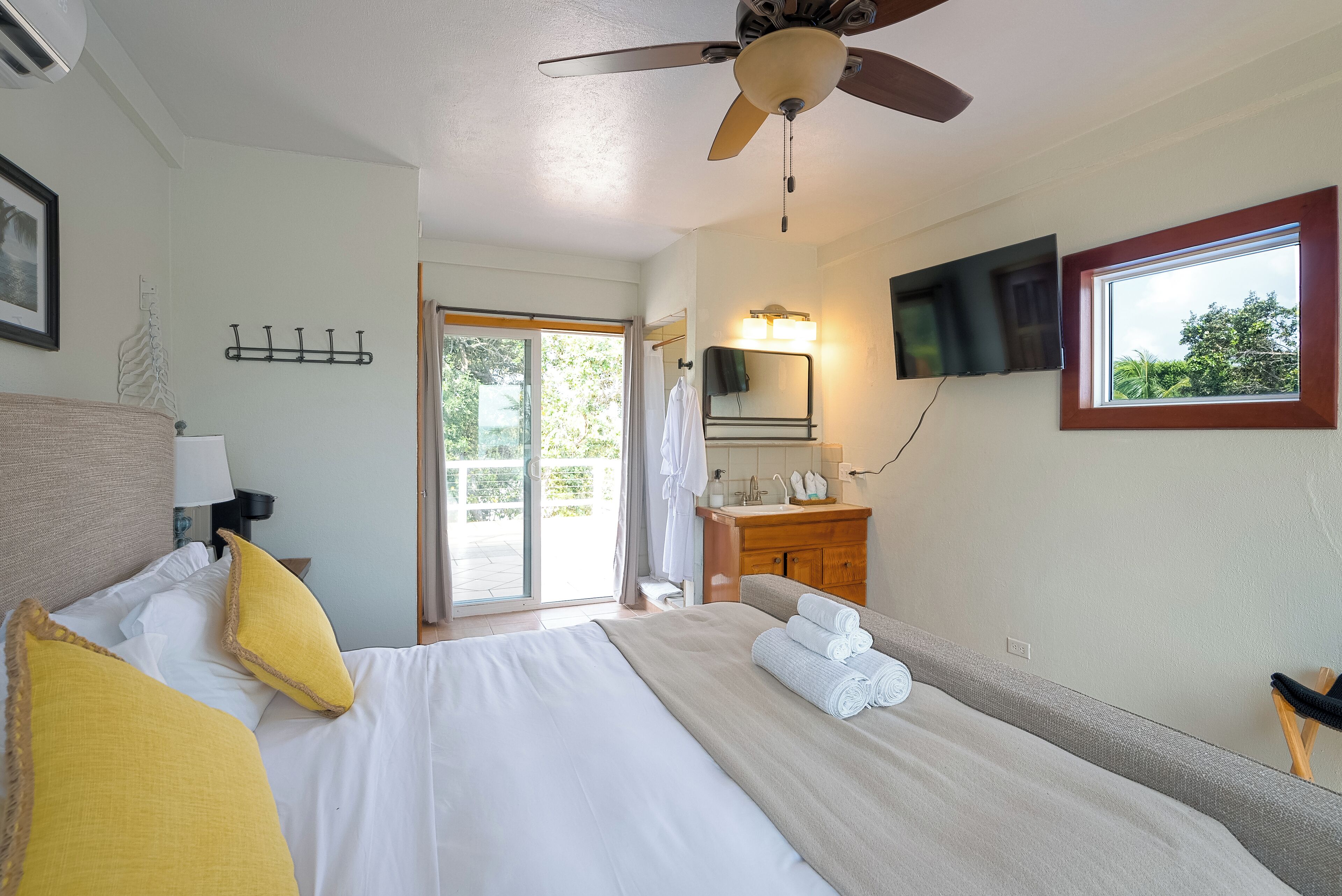 Une chambre d’hôtel équipée d’un lit, d’un ventilateur de plafond, d’une télévision, d’une salle de bain avec un lavabo et un miroir, et d’une fenêtre donnant sur un paysage verdoyant.