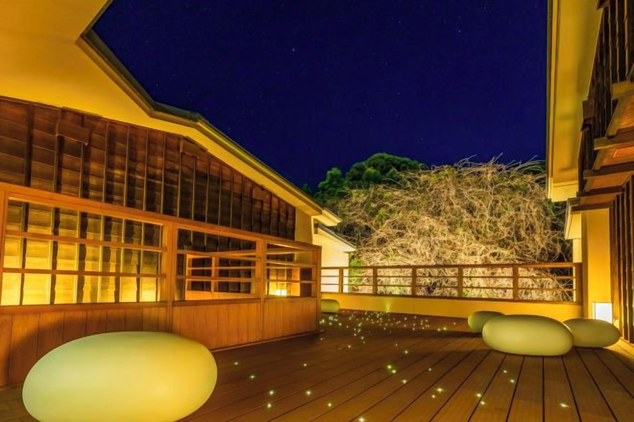 Un bâtiment traditionnel japonais doté d’une terrasse en bois, de grandes fenêtres et offrant une vue sur un arbre.