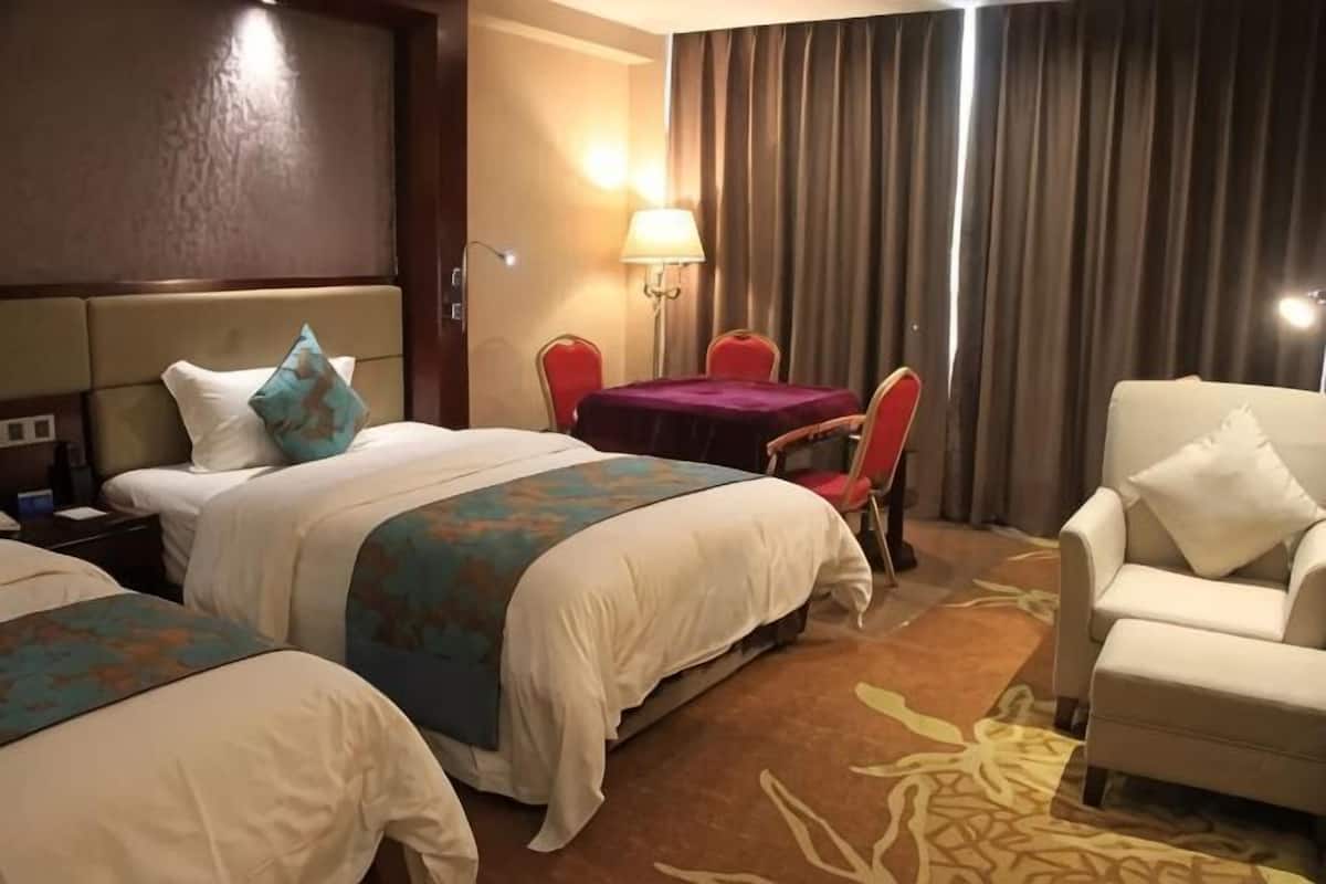 A hotel room with two beds, a desk, a chair, a lamp, and a view of the outside.