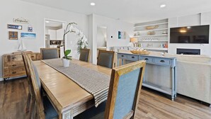 A modern dining area with a wooden table, chairs, and a chandelier. There is a kitchen area with a microwave and a TV on the wall.