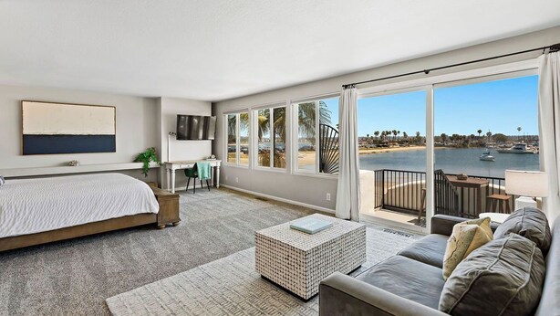 A spacious bedroom with a large bed, a seating area with a sofa and a coffee table, and a balcony with a view of the sea and boats.