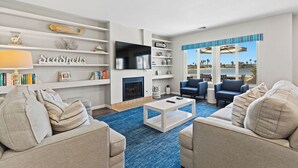 A modern living room with a fireplace, a flat-screen TV, and a view of the beach through large windows.