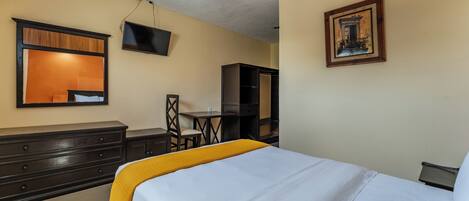Quarto de hotel com cama, escrivaninha, cadeira, guarda-roupa, televisão e um quadro na parede.