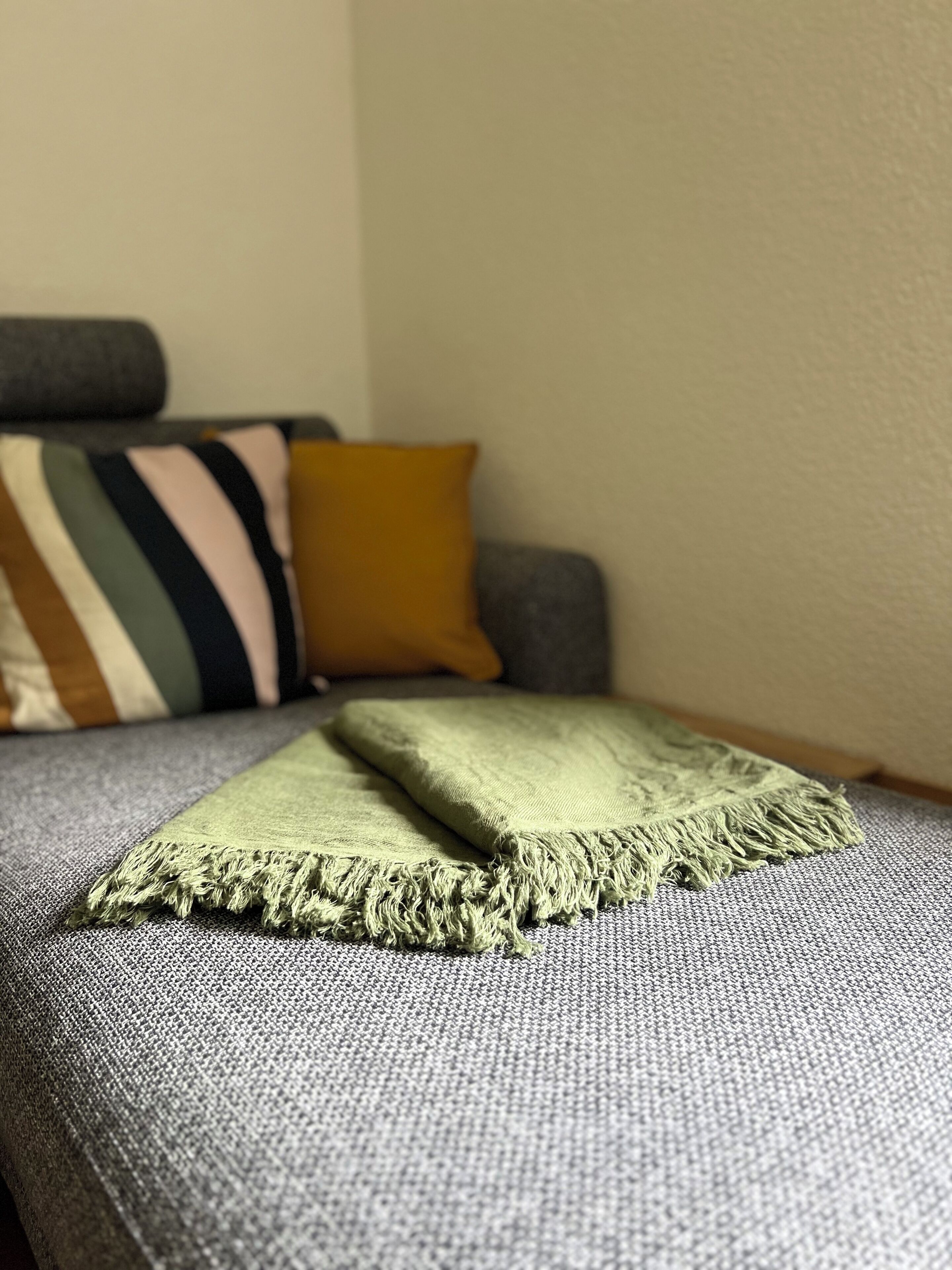 Een bed met een grijs geweven dekbedovertrek, een groen deken met franjes, en een gestreept kussen.
