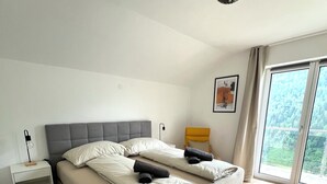 A modern bedroom with a large bed, a bedside table, a nightstand, a window with a view of greenery, and a ceiling light fixture.