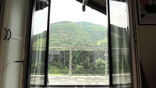 A room with a large sliding glass door leading to a balcony with a view of green hills.
