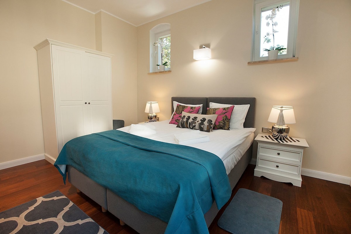 Une chambre à coucher comprenant un lit, des tables de chevet, une commode, un banc, une armoire et une fenêtre agrémentée d’une plante orchidée.