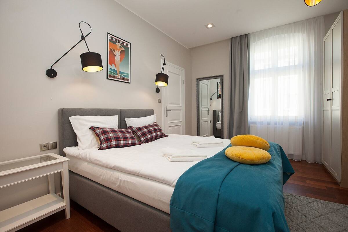 Une chambre moderne avec une tête de lit grise, du linge de lit blanc, un plaid turquoise et des coussins jaunes. On y trouve deux lampes suspendues noires, une affiche encadrée au mur et un grand miroir au-dessus de la porte.