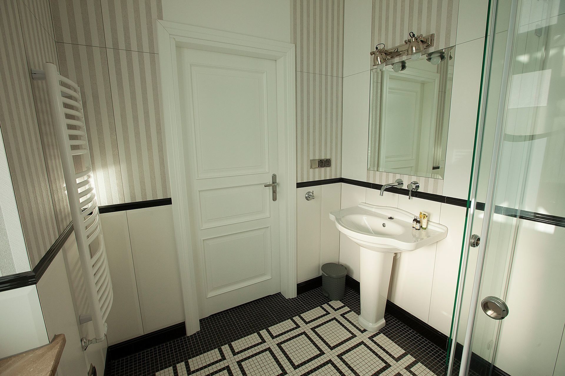 Un baño con una puerta blanca, cabina de ducha de vidrio, lavamanos blanco y piso estampado.