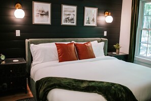 Un lit avec des oreillers blancs et bruns, un plaid vert et une tête de lit. On y voit également des cadres photos fixés au mur et deux appliques murales.
