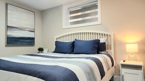 A neatly arranged bedroom with a large bed, a bedside table with a lamp, a window with blinds, and a painting on the wall.