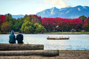 Two people sitting on a wooden bench by a lake with a dock and birds, surrounded by trees with autumn foliage and mountains in the background.