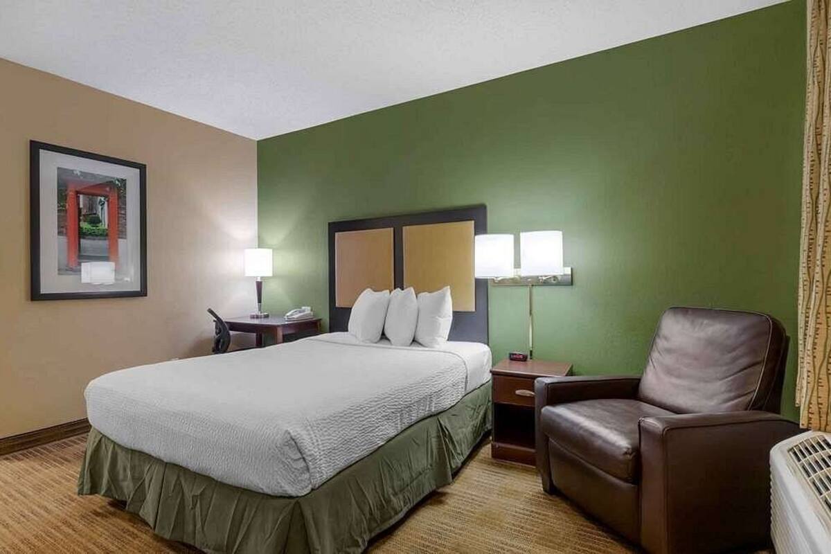 A hotel room with a bed, a desk, a chair, and a lamp.