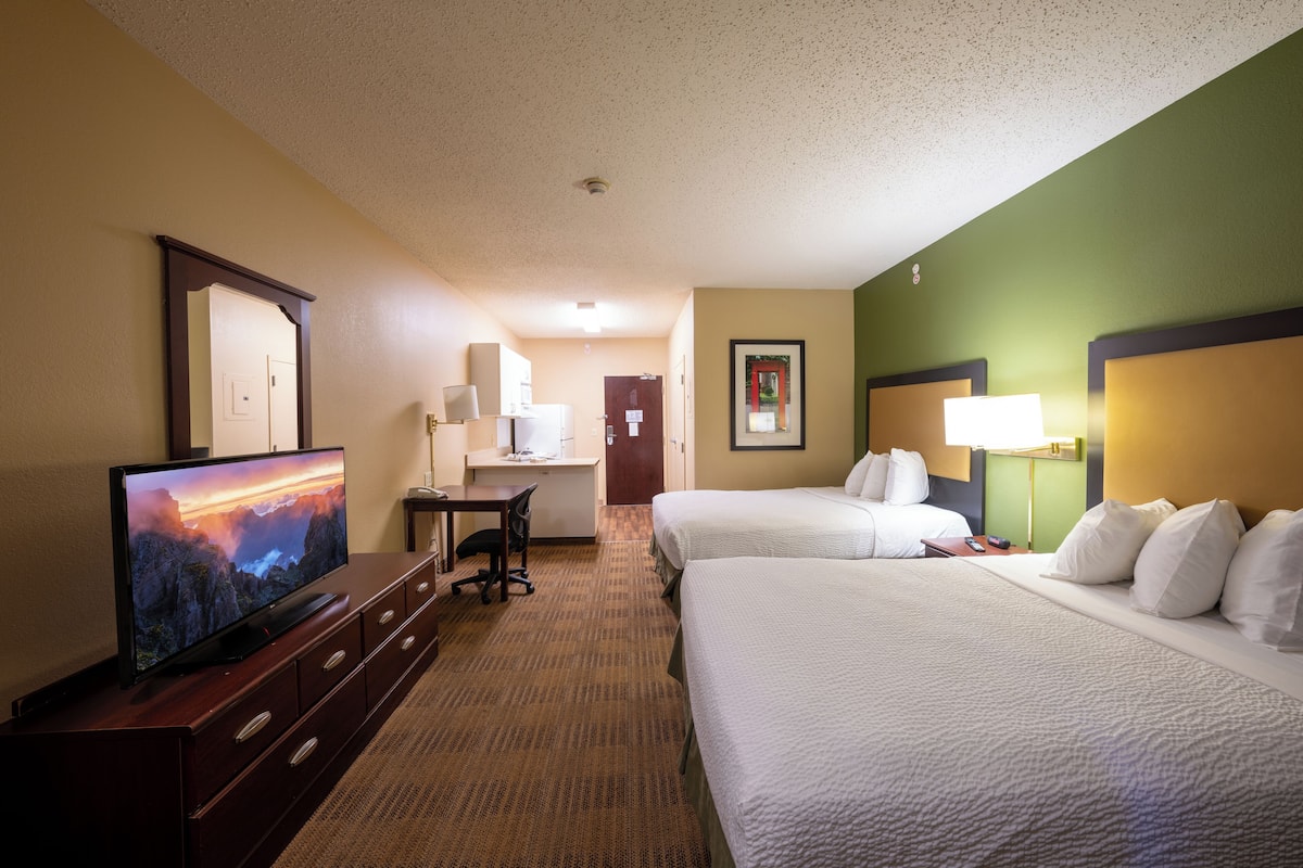 A hotel room with two beds, a desk with a chair, a television, and a bathroom visible in the background.