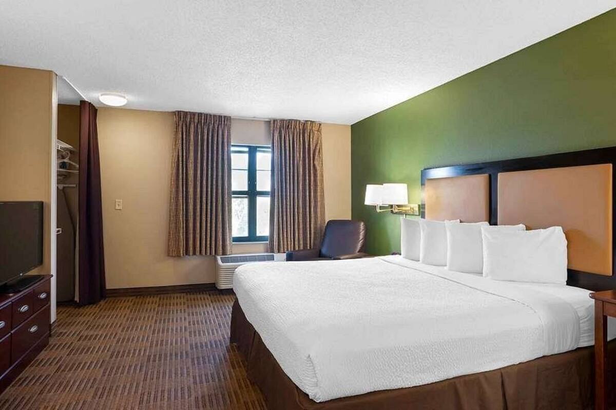 A hotel room with a large bed, a TV, a chair, and a window with curtains.
