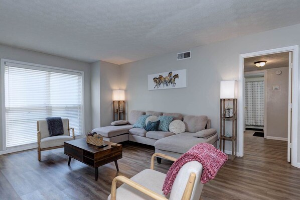 A living room with a sofa, two armchairs, a coffee table, a lamp, and a painting on the wall. - Graduation Basecamp - Family 2BR Near Ft Benning (Columbus)