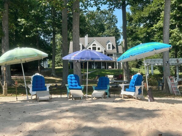 A beach area with four blue lounge chairs under umbrellas, a house in the background, and a pergola with a blue umbrella.