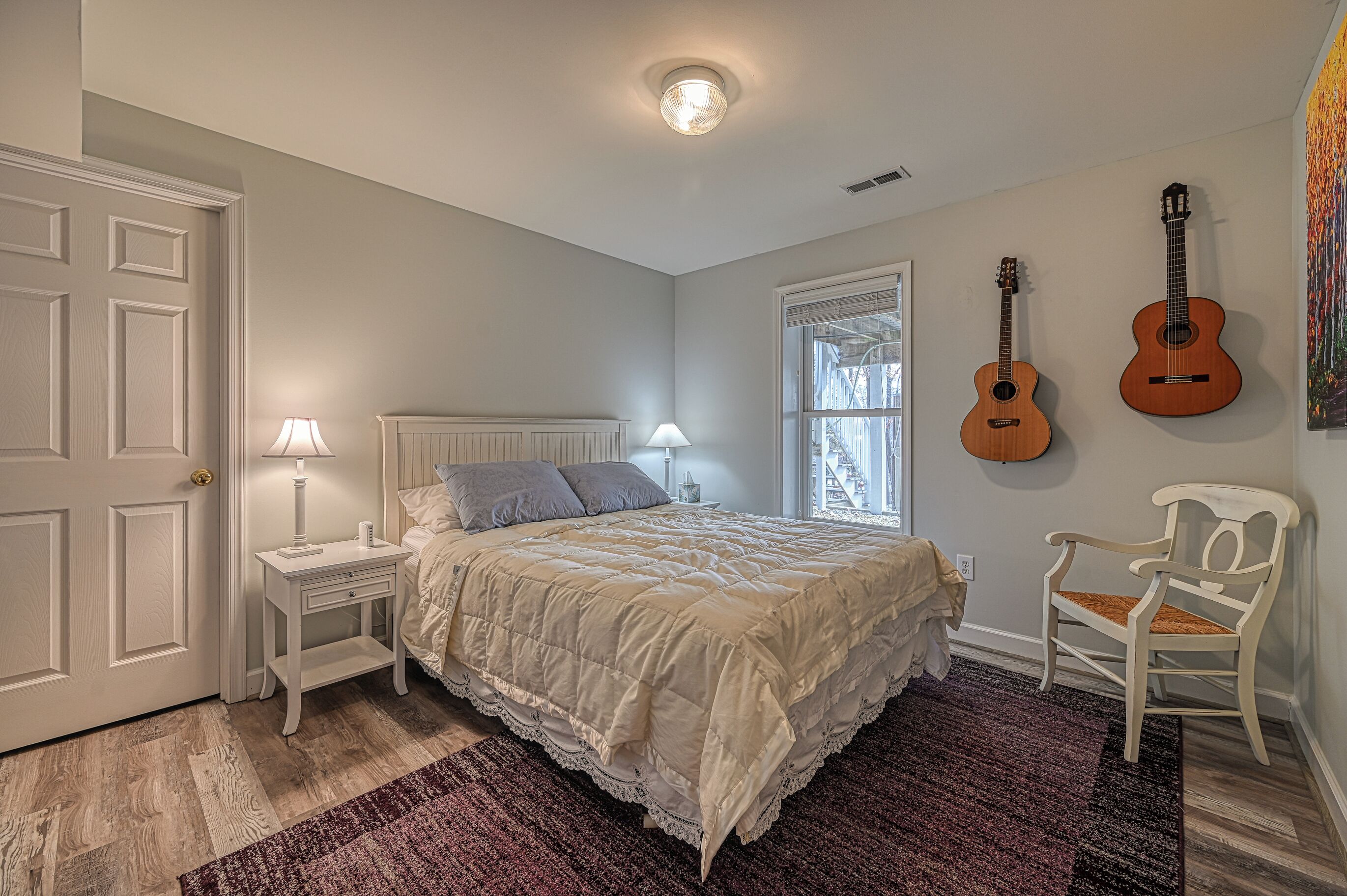 A bedroom with a bed, nightstand, lamp, two guitars, and a chair.