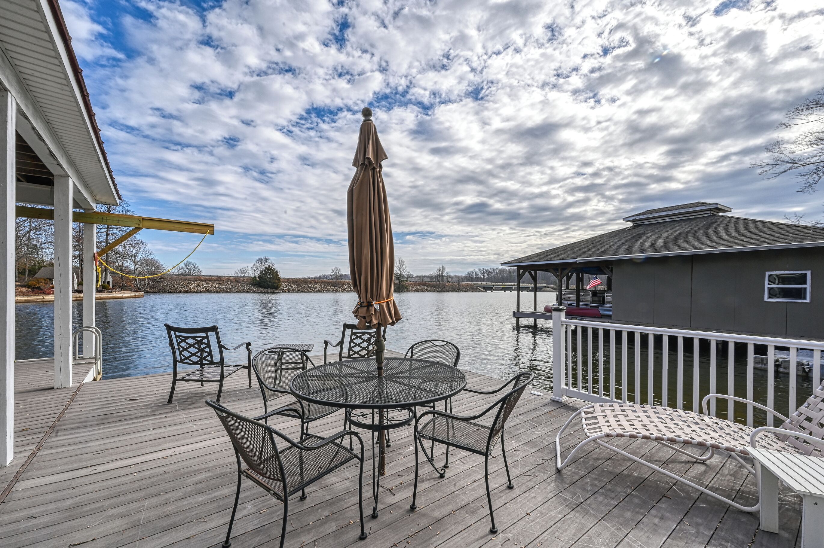 A wooden deck with a round table and chairs, a covered umbrella, and a white railing overlooking a body of water.