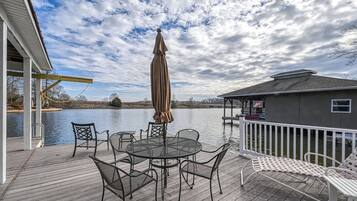 A wooden deck with a round table and chairs, a covered umbrella, and a white railing overlooking a body of water.