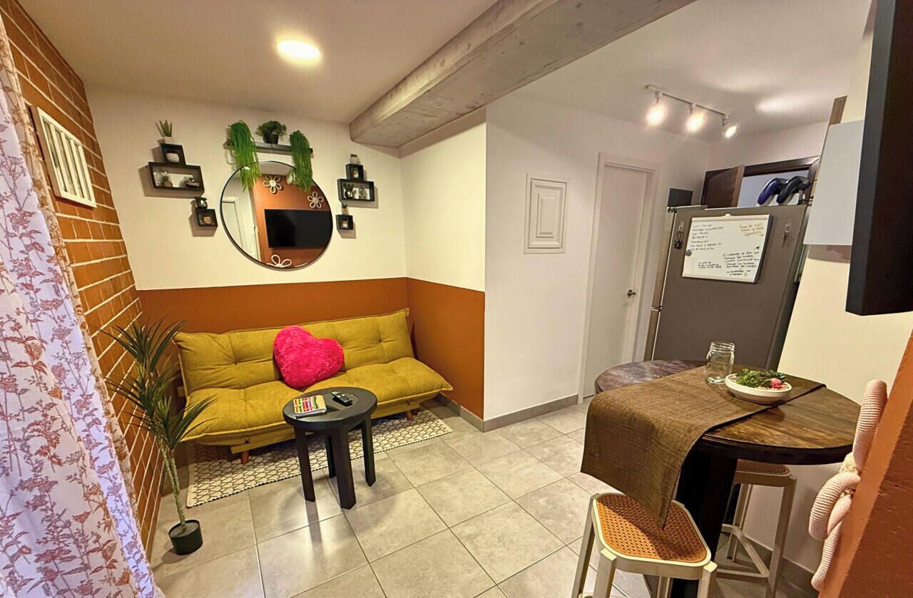A compact living space with a sofa, a small table, a dining table, a refrigerator, and a mirror on the wall.