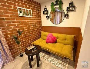 Un salon avec un mur en briques, un canapé agrémenté d’un coussin en forme de cœur, une petite table avec une télécommande et un miroir décoré de plantes.