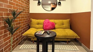 Um quarto com parede de tijolos, sofá amarelo, espelho redondo e uma pequena mesa com controle remoto.