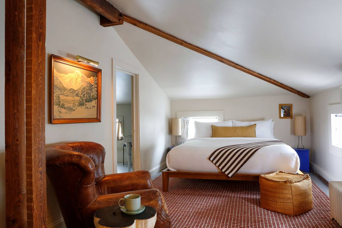 Een slaapkamer met een groot bed, een houten stoel, een klein tafeltje met een kopje, en een schilderij aan de muur.