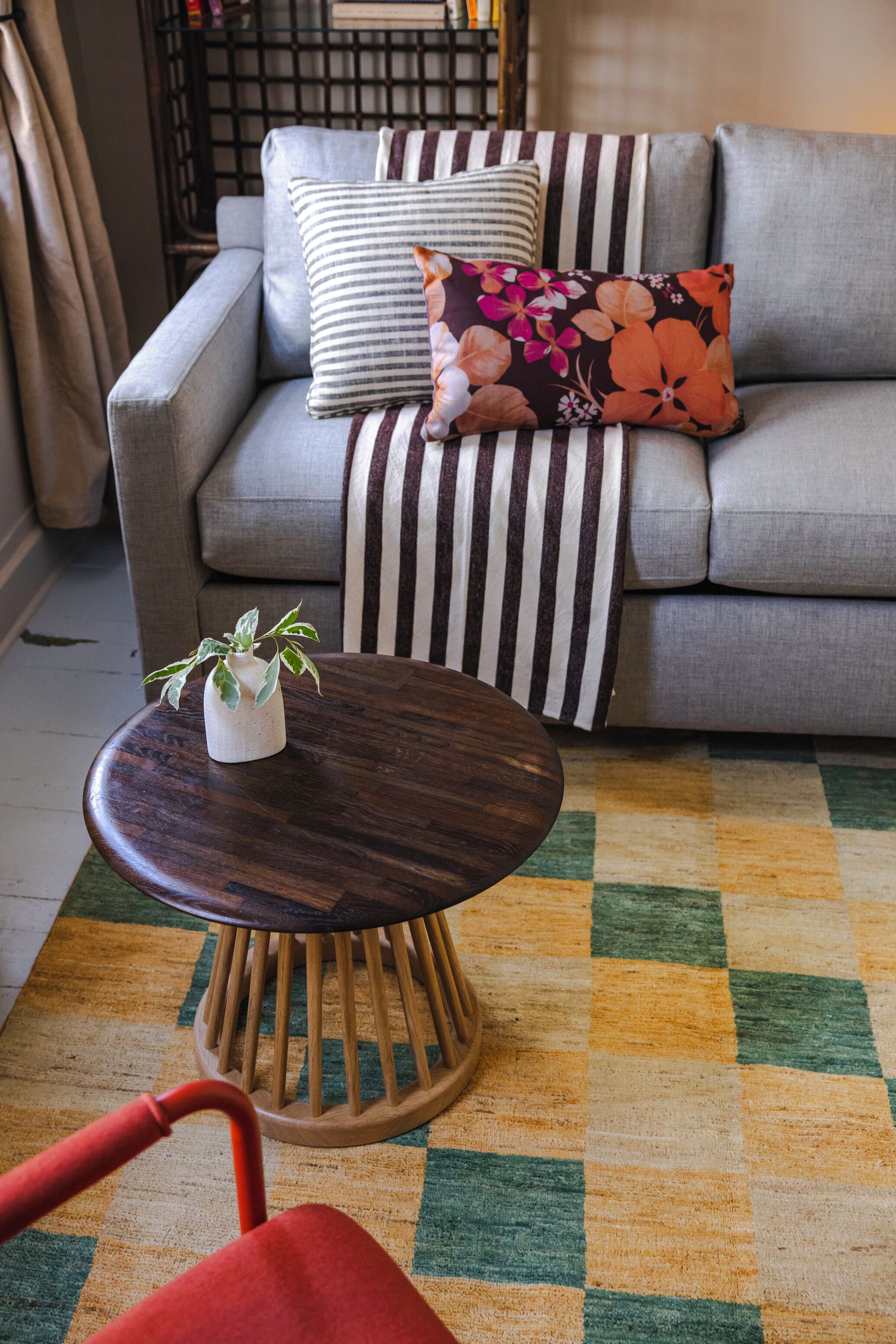 A living room with a grey sofa, striped pillows, a small round wooden table with a plant, and a checkered rug.
