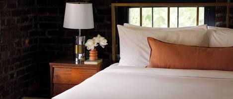 A neatly made bed with white linens and a single brown pillow, a bedside table with a lamp and a vase of flowers, and a brick wall in the background.