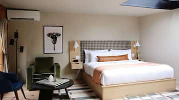 A hotel room with a large bed, a green armchair, a small table with a book, and a framed picture on the wall.