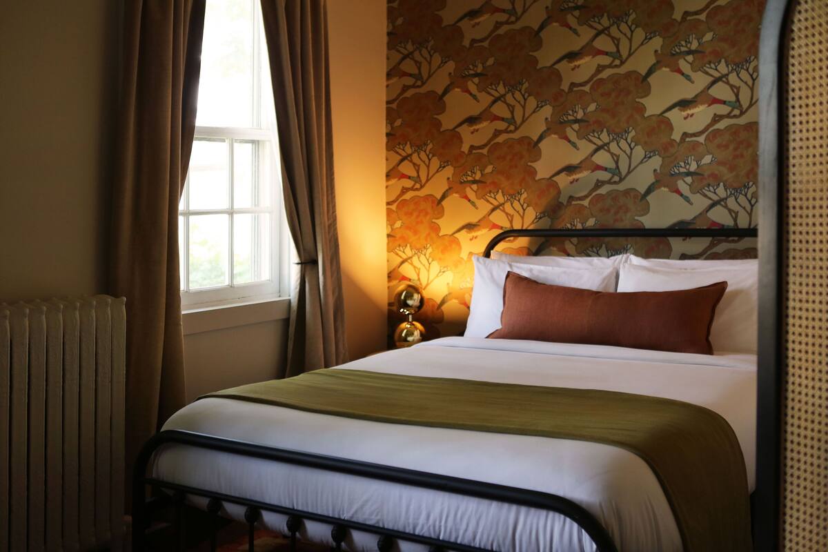 Een net opgemaakt bed met een wit- en groen beddekleed, een bruin kussen en een bloemen behang.