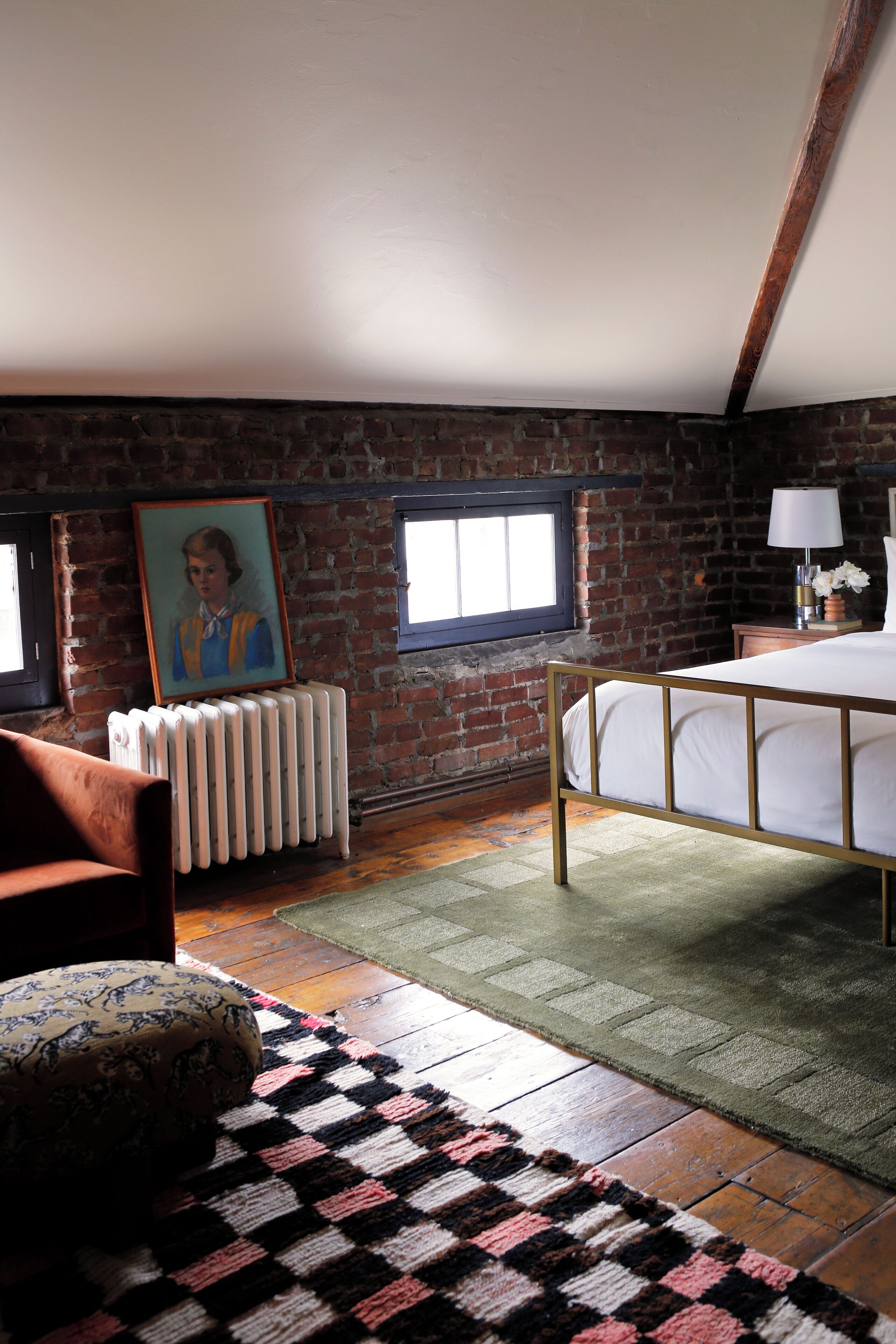 A bedroom with a brick wall, a radiator, a bed, a window, and a painting on the wall.