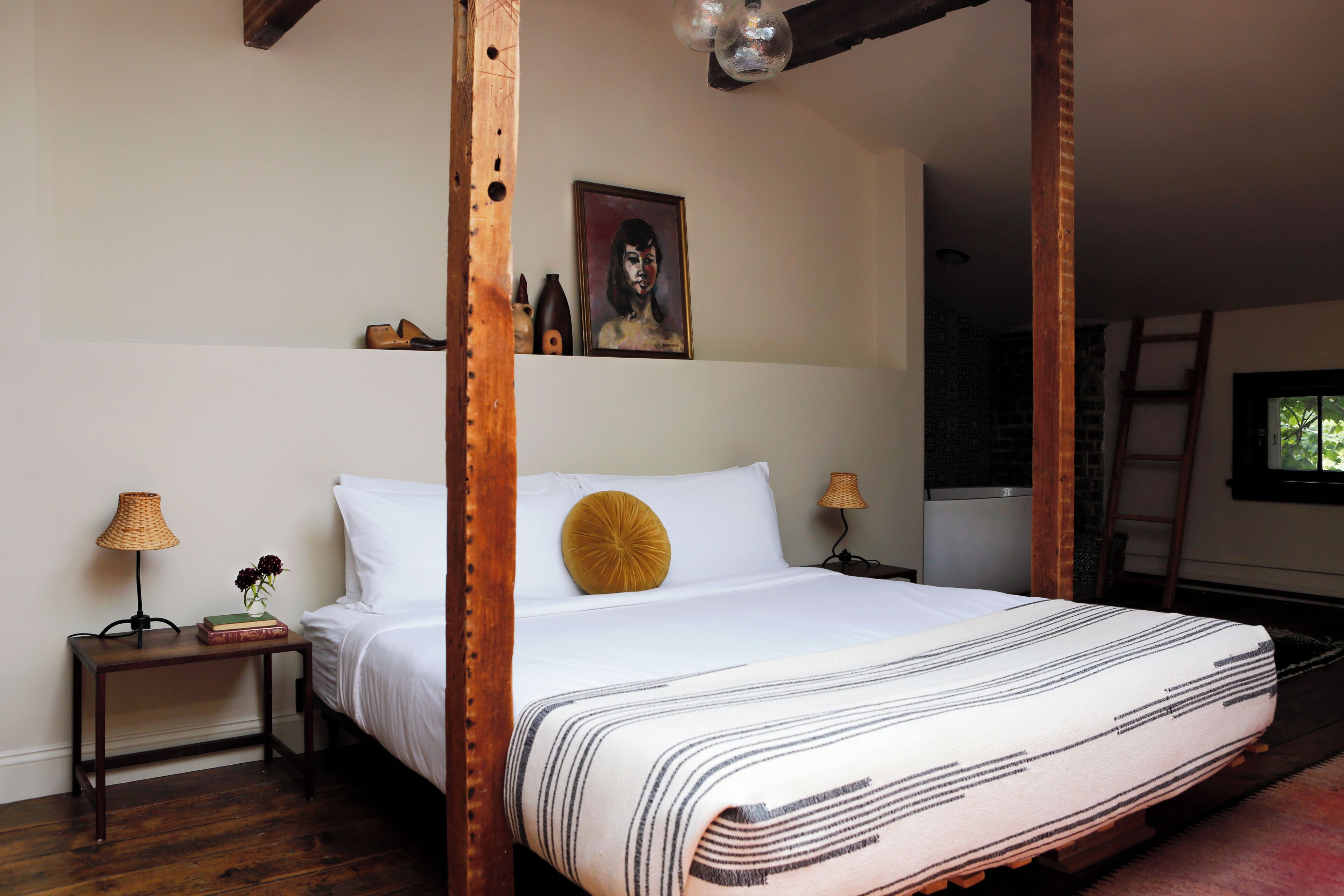 A bedroom with a four-poster bed, a painting on the wall, a small table with a lamp, and a window with a view of trees.
