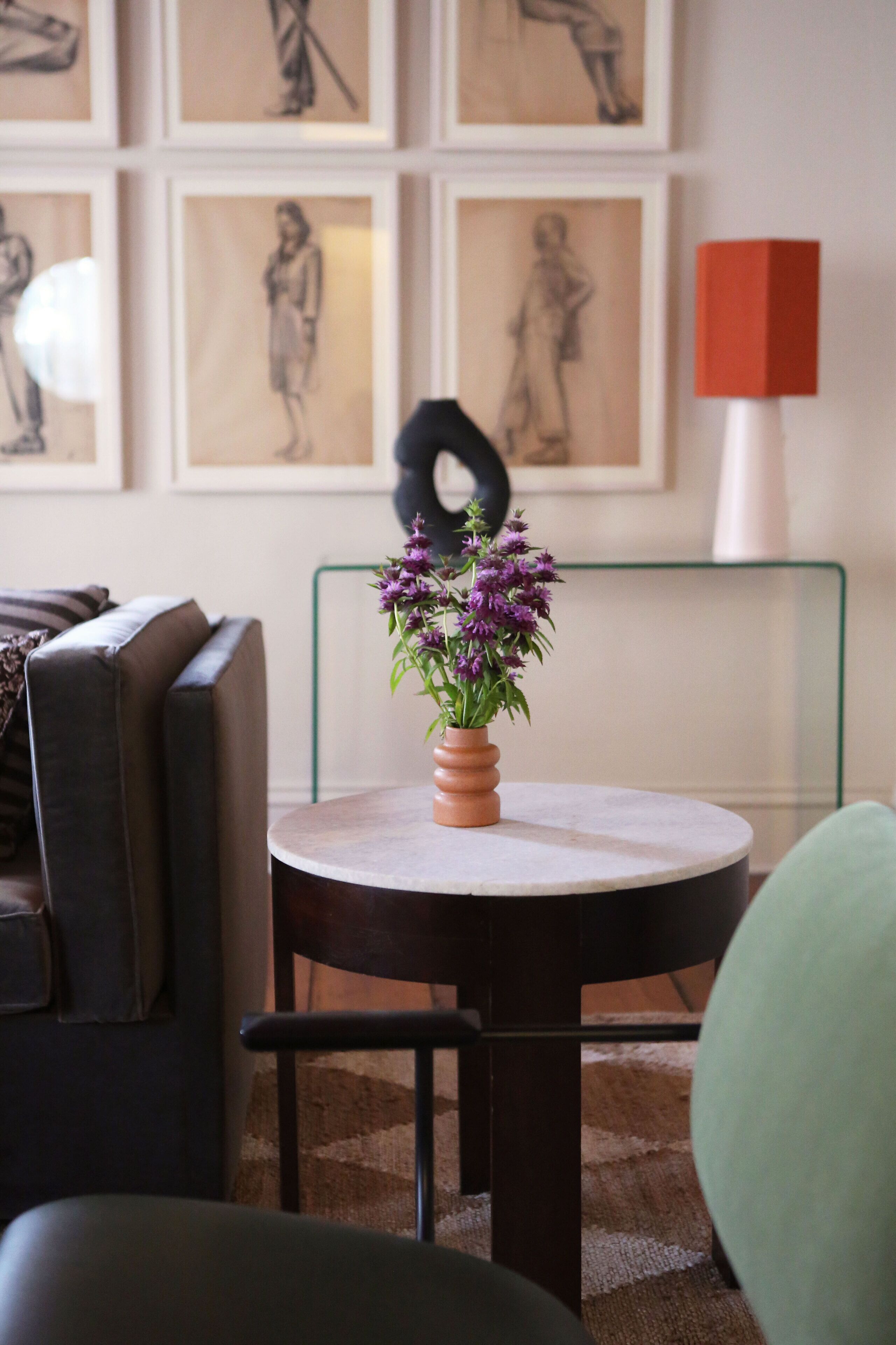 Uma mesa redonda com um vaso de flores roxas, uma cadeira preta e uma cadeira verde em um quarto com um sofá e obras de arte emolduradas na parede.