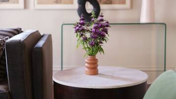 Uma mesa redonda com um vaso de flores roxas, uma cadeira preta e uma cadeira verde em um quarto com um sofá e obras de arte emolduradas na parede.