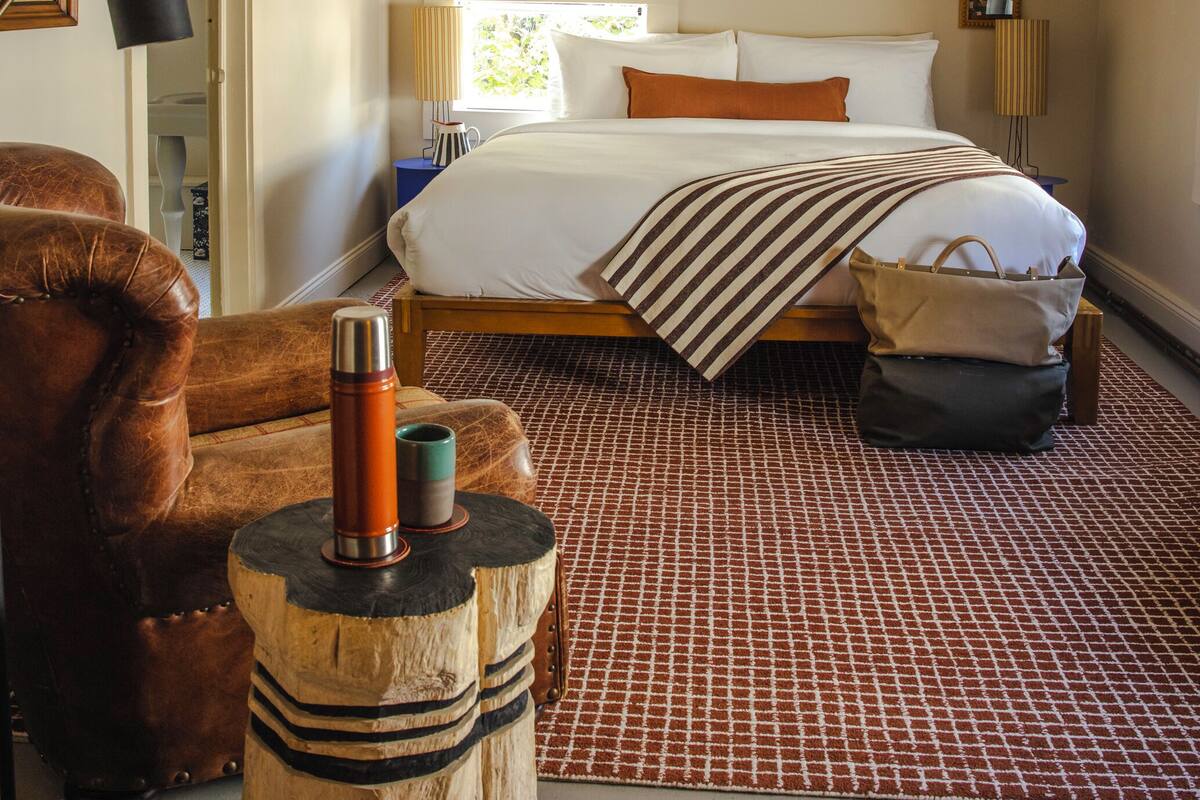 Een slaapkamer met een groot bed, een houten hoofdbord, een tapijt met rood-witte patronen en een leren fauteuil.