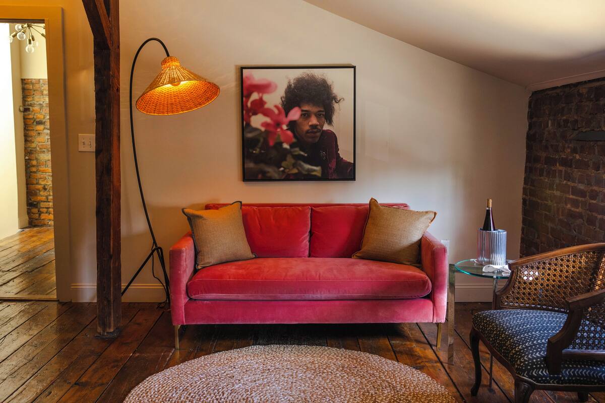 Een rode bank met twee kussens, een rieten stoel, een rond vloerkleed, een wandlamp en een ingelijst schilderij aan de muur.