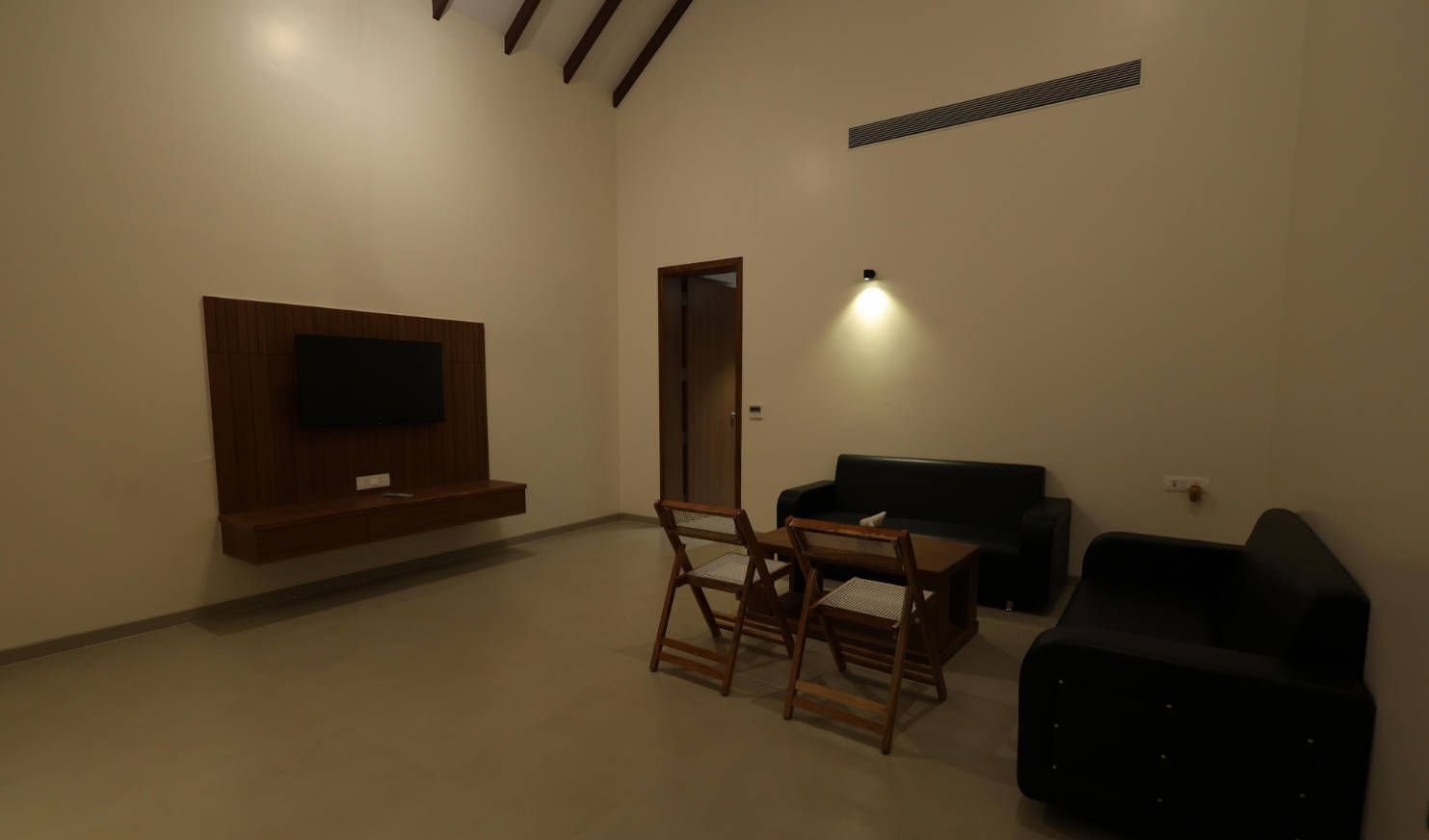 A living room with a flat-screen TV mounted on the wall, a dining table with chairs, a black sofa, and a black armchair.