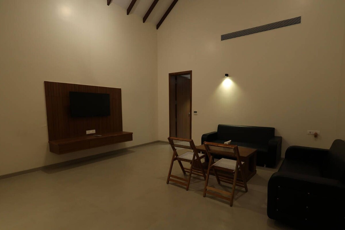 A living room with a flat-screen TV mounted on the wall, a dining table with chairs, a black sofa, and a black armchair.