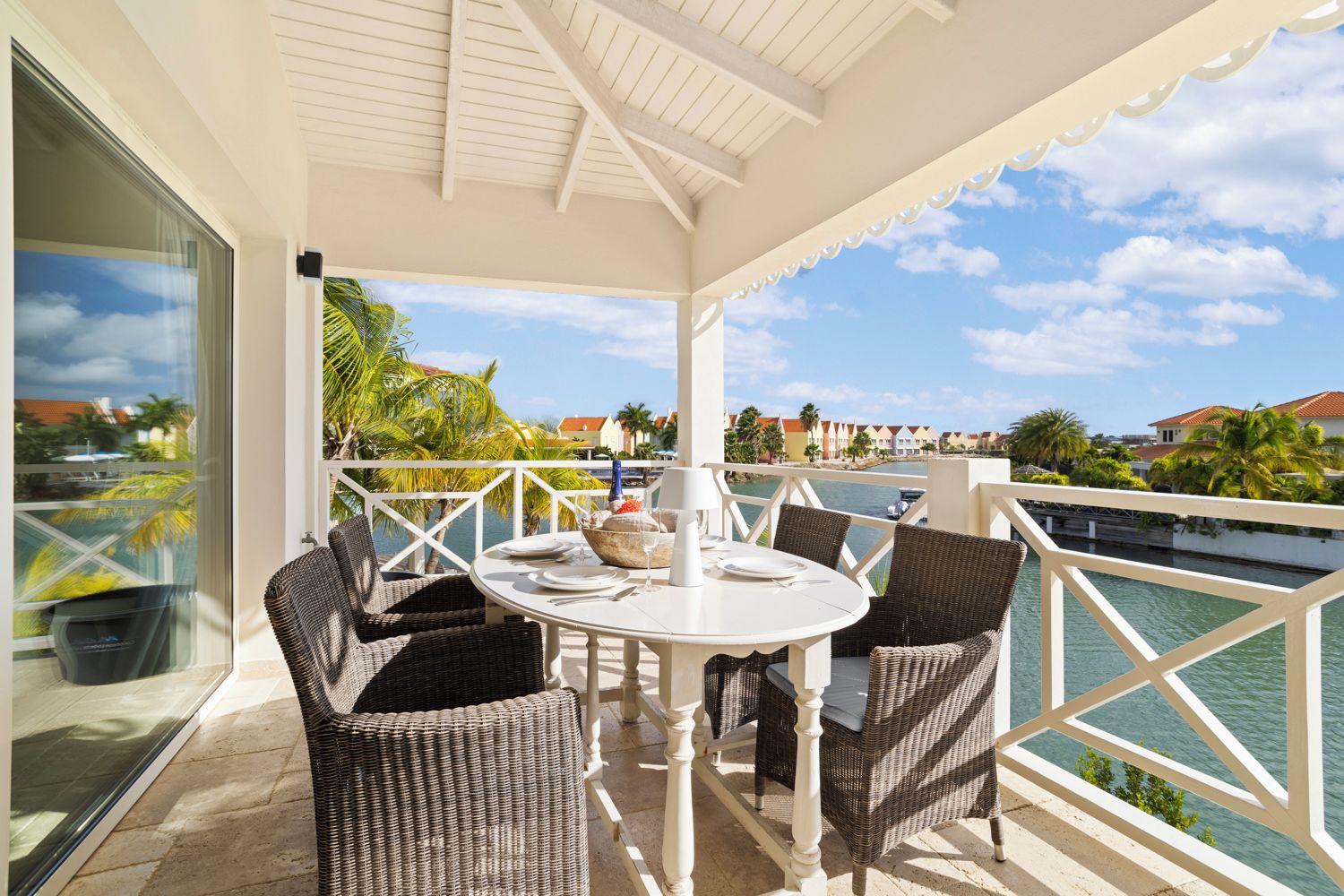 A covered outdoor dining area with wicker furniture and a table set for two, overlooking a body of water and a row of buildings.