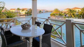 A covered outdoor seating area with a table and chairs, overlooking a body of water and a row of buildings.