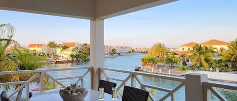 A covered outdoor seating area with a table and chairs, overlooking a body of water and a row of buildings.